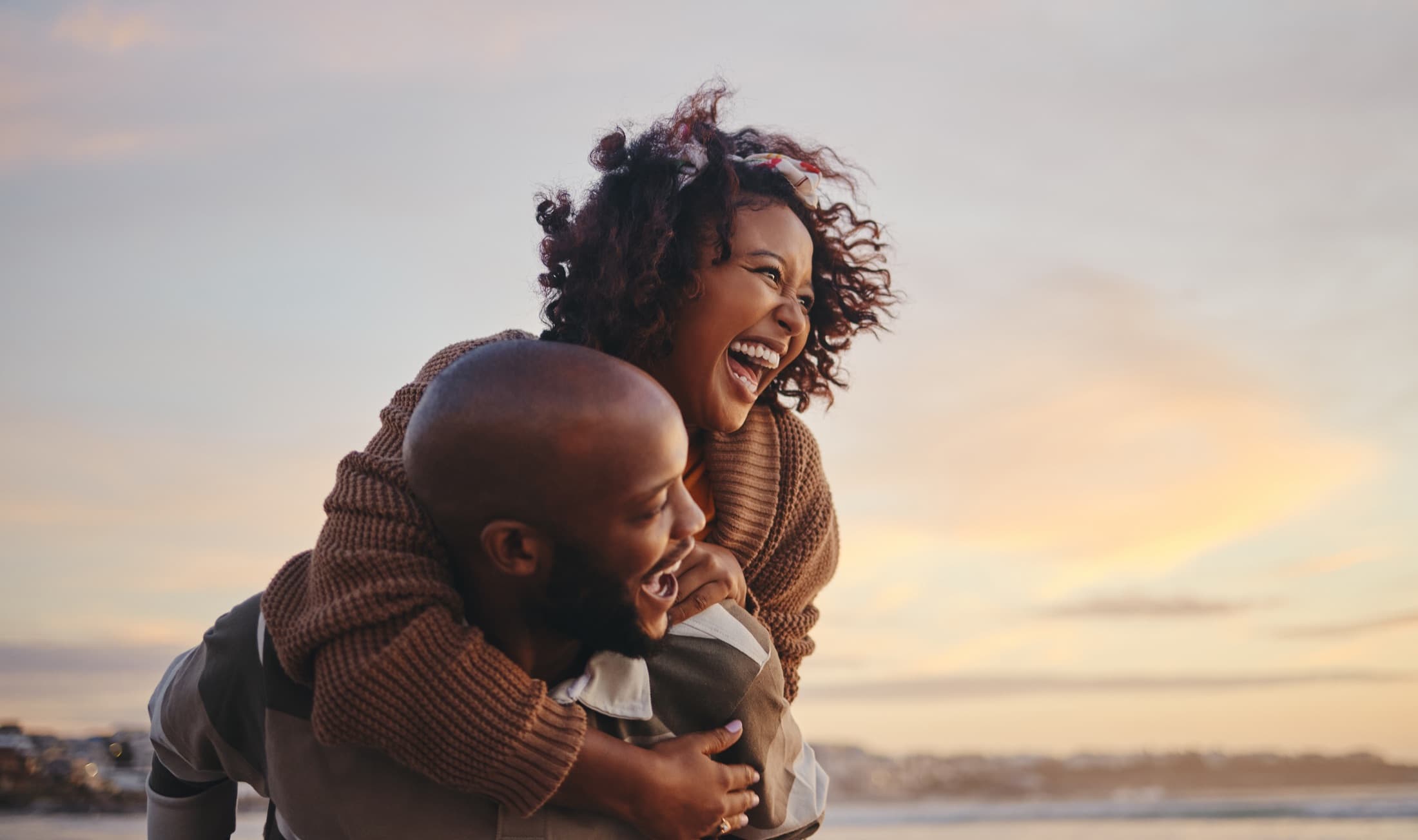 A woman laughing as she rides piggyback on her partner on a beach at sunset