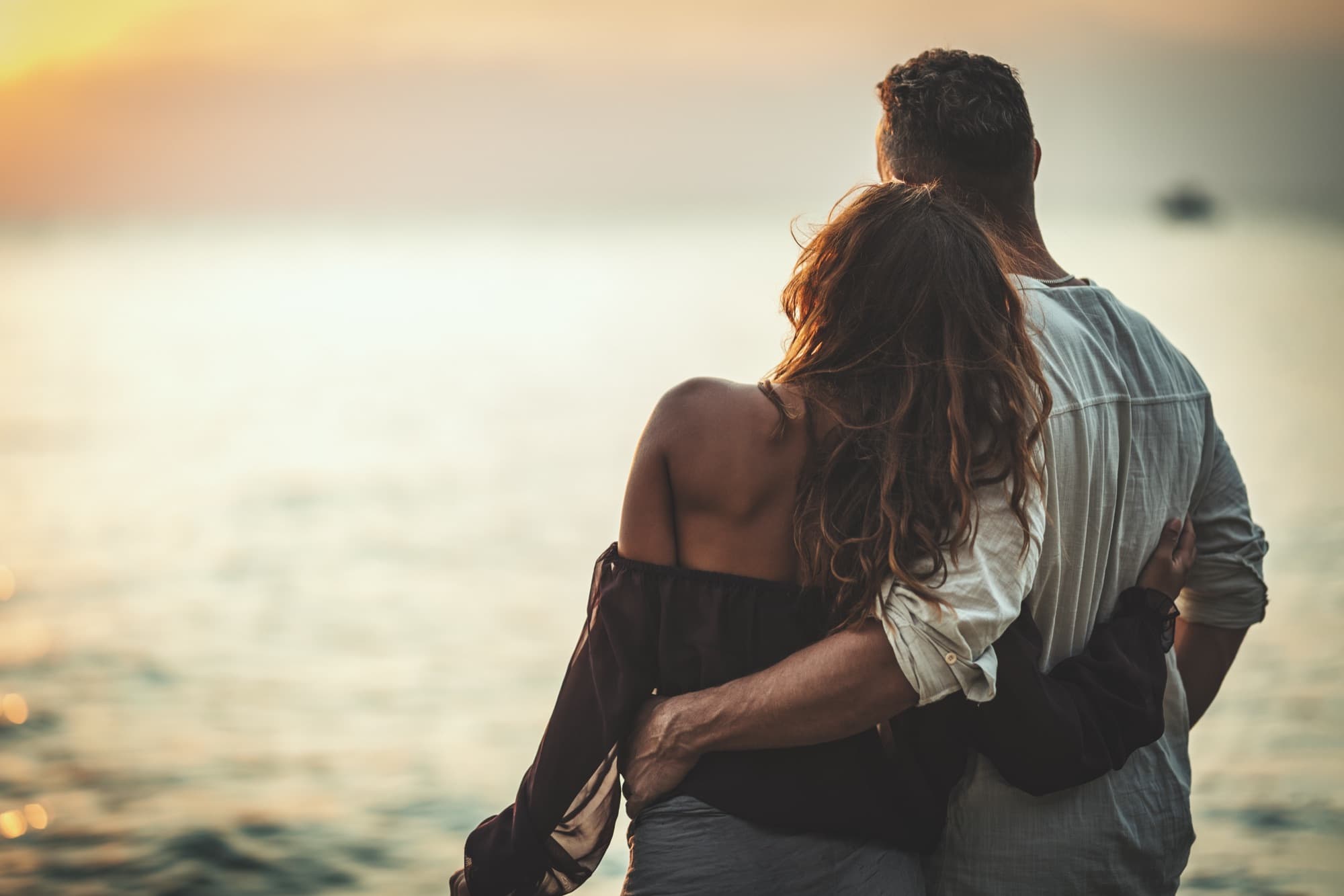 A couple seen from behind, arms around each other, looking out over the sea at sunset