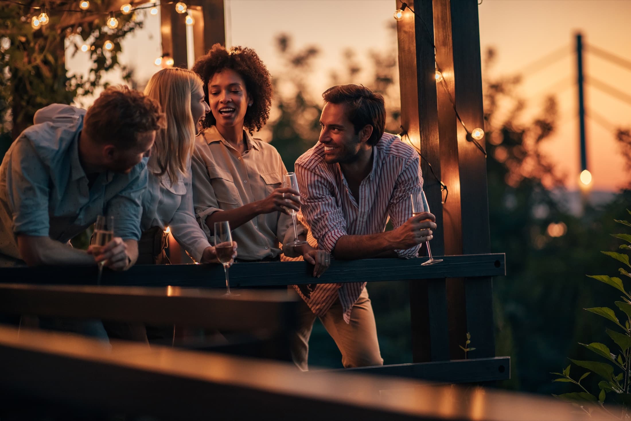 Four friends laughing with glasses of wine at a terrace railing under string lights at dusk