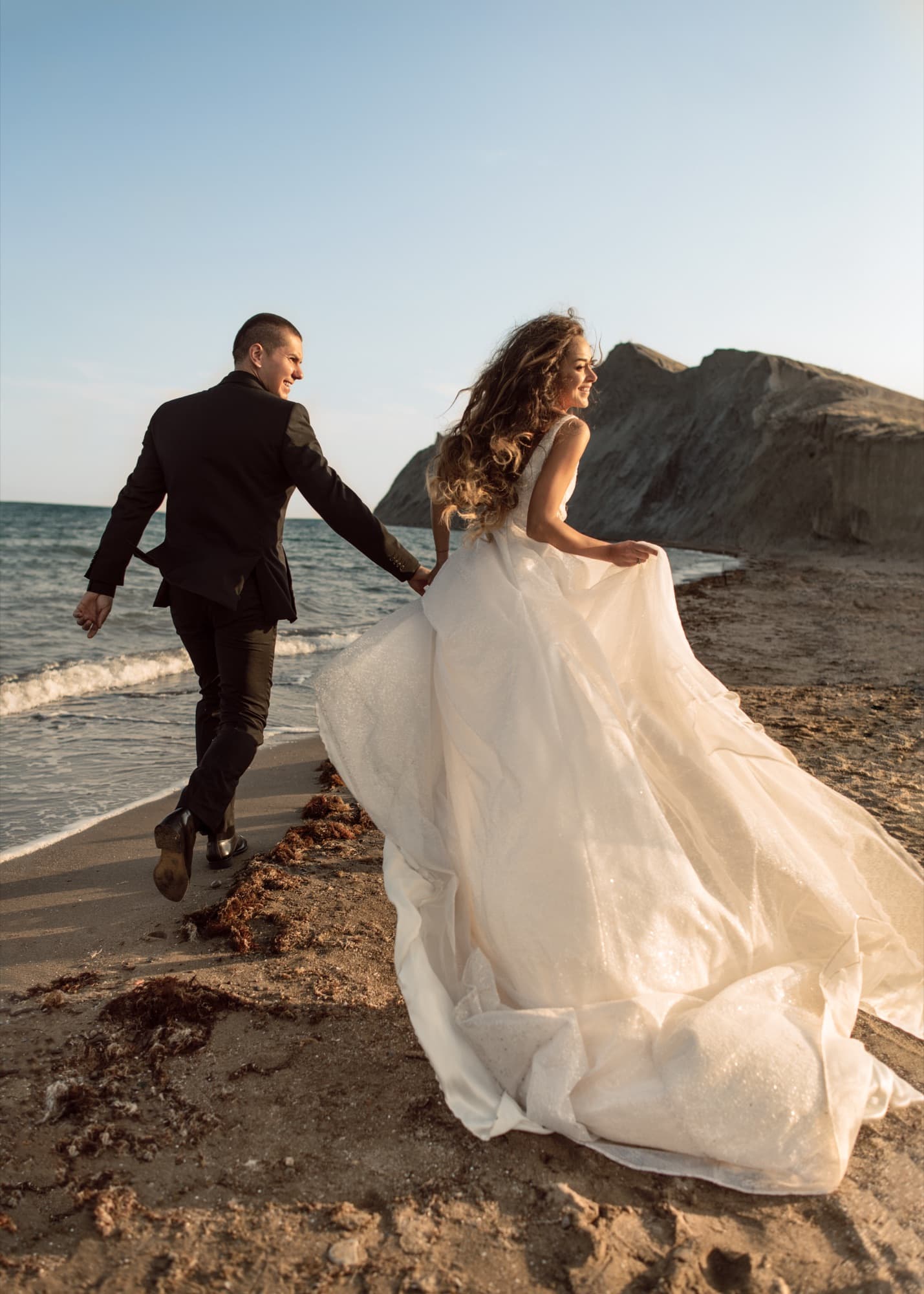 A bride and groom running hand in hand along a rocky beach, her gown trailing behind